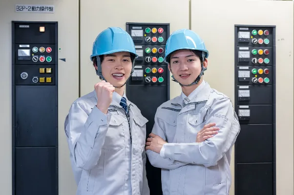 制御盤の前でガッツポーズをする作業服姿の女性作業員2人